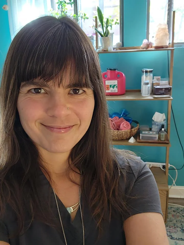 Briana Sabaj in her treatment room, smiling, with herbs and supplies on the shelves behind her